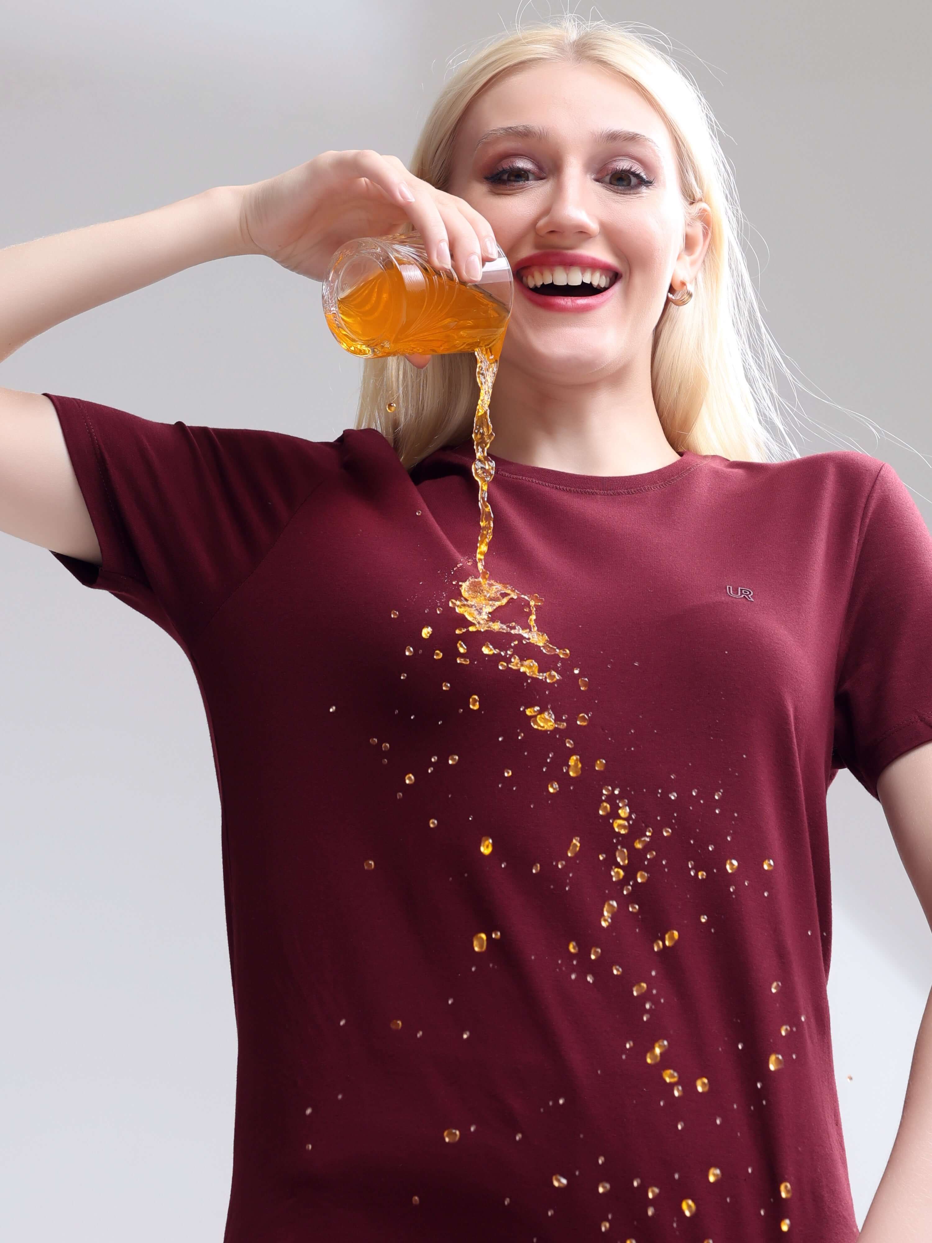 Woman wearing cranberry stain-proof, odor-resistant round-neck Turms T-shirt demonstrating spill resistance with liquid pour.