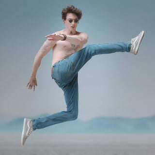 Man in sunglasses performing a dynamic leap, showcasing stylish jeans and athletic shoes against a soft blue background.