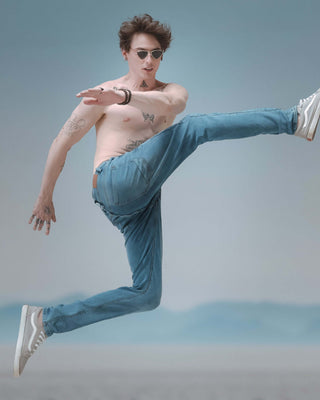 Man in sunglasses performing a dynamic leap, showcasing stylish jeans and athletic shoes against a soft blue background.