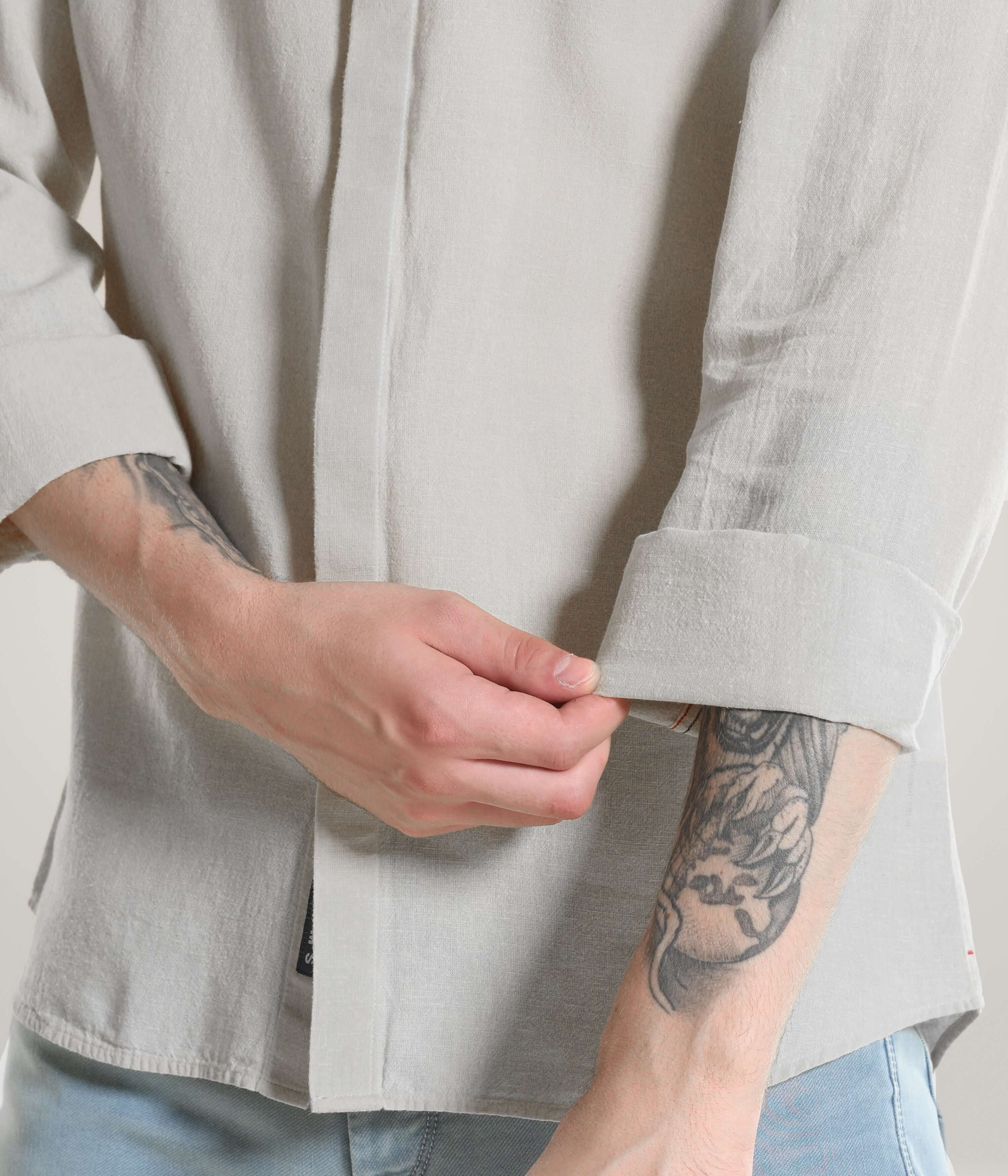 Person rolling up the sleeve of a Storm Grey Cotton/Linen Turms shirt showcasing meticulous stitching and tattooed arm.