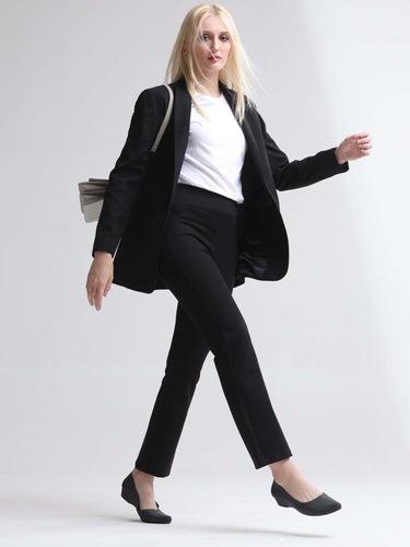 Woman in black blazer and pants, white t-shirt, black flats, carrying a handbag, walking confidently against a minimalist background.