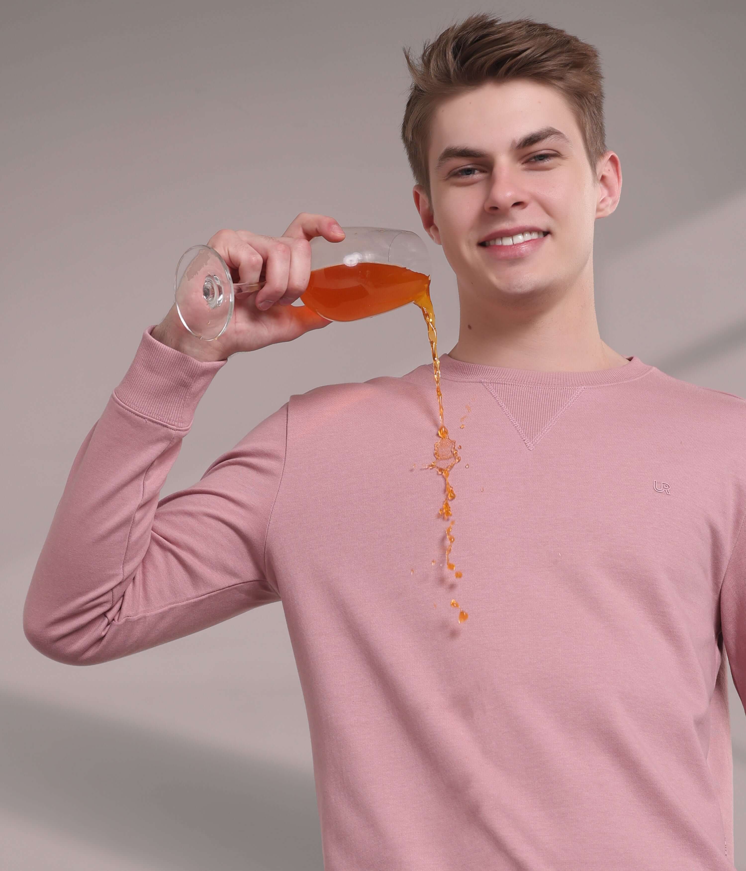 Young man in a pink sweatshirt demonstrating its stain-proof features by pouring juice, showcasing style and functionality.