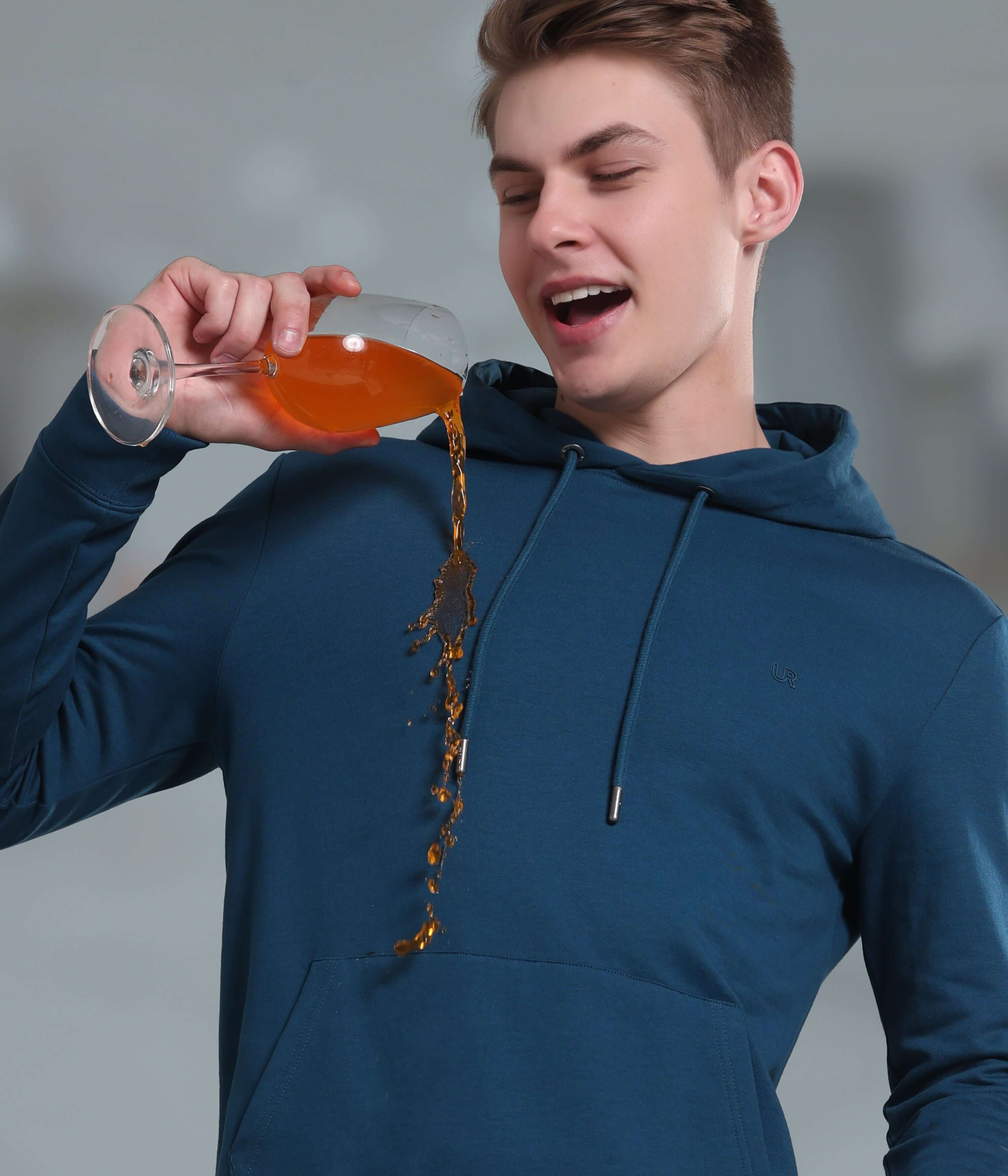 Young man wearing a blue hooded sweatshirt, demonstrating its waterproof and stain-proof features while pouring a drink.