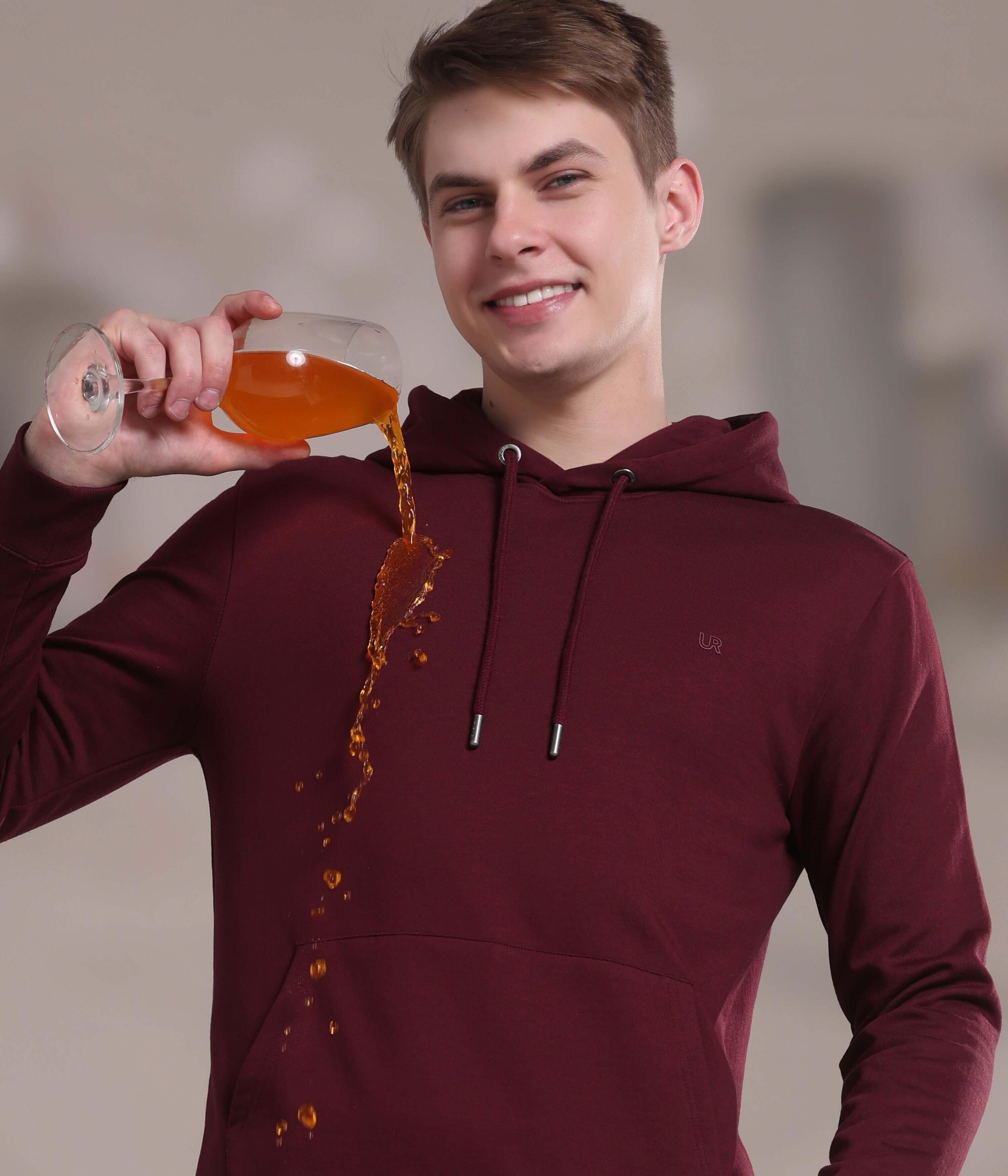 Model in a burgundy hooded sweatshirt demonstrating its waterproof, stain-proof features by spilling a drink.