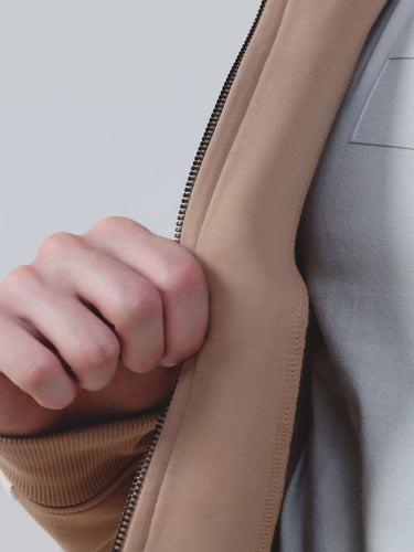 Close-up of a hand holding the zipper of the Turms Intelligent Cord Set jacket in sandy color.