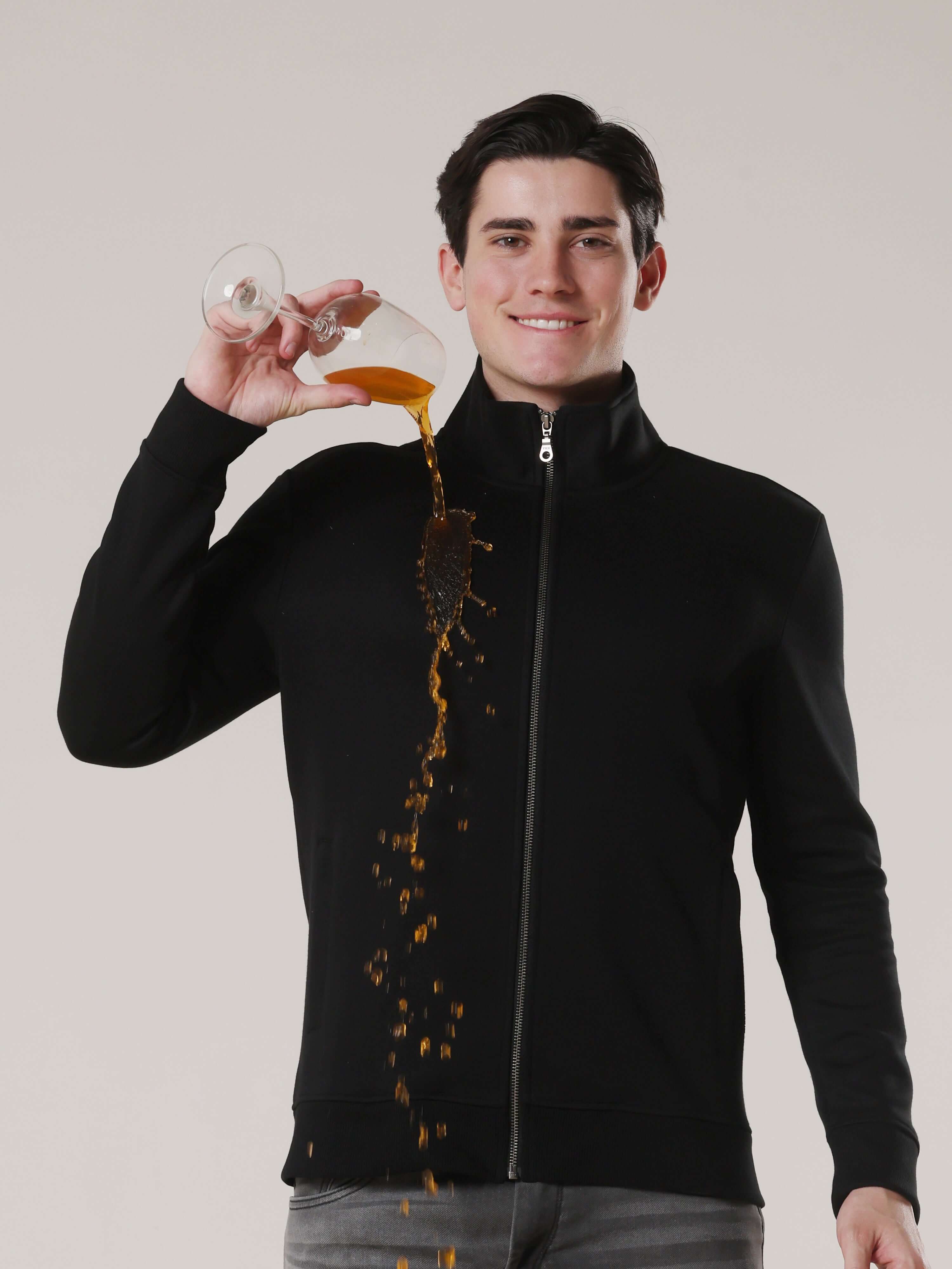 Model wearing a black jacket, showcasing its water-repellent feature while liquid spills from a glass.
