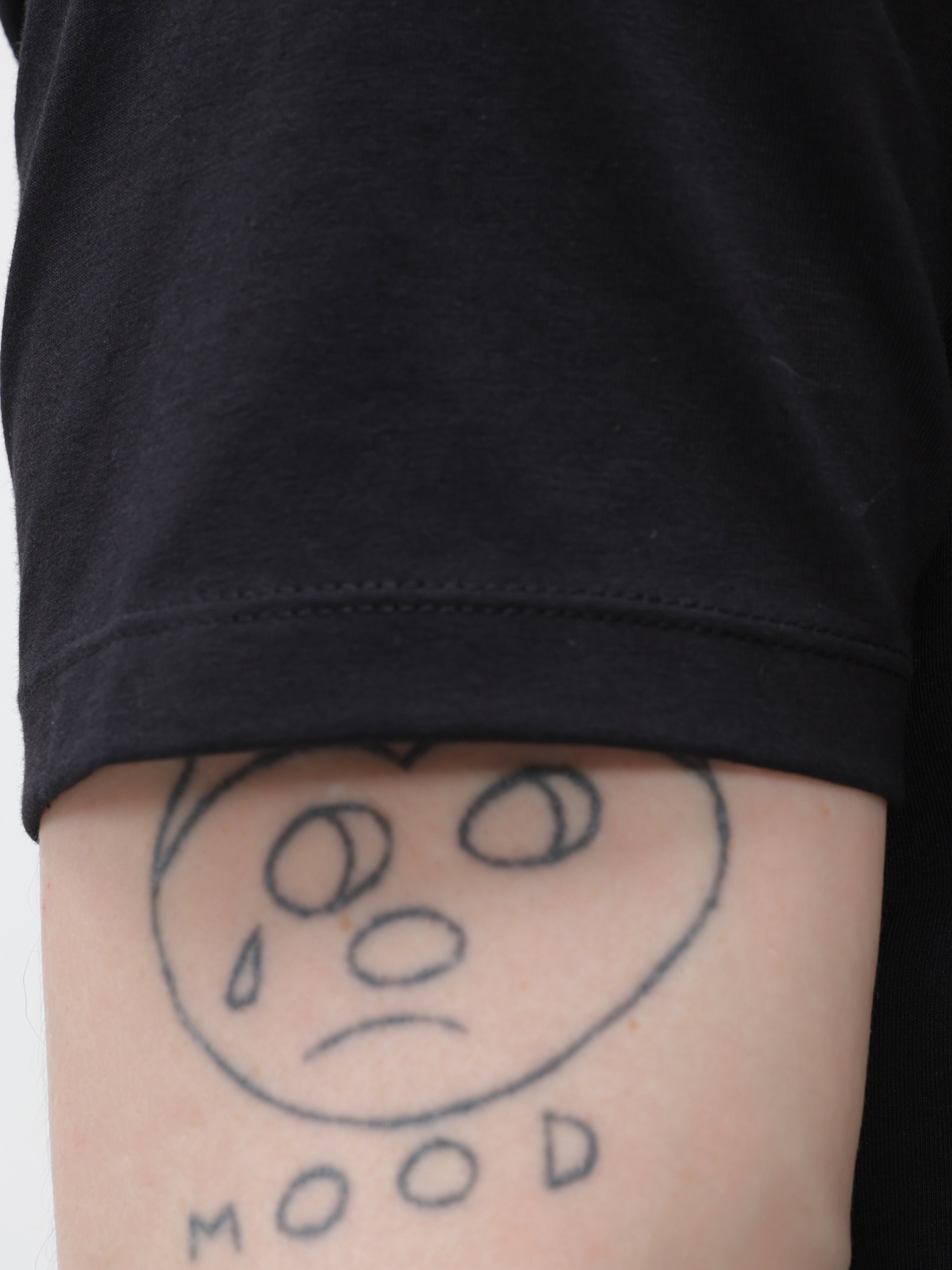 Close-up of arm wearing a black round-neck t-shirt with a "Mood" tattoo featuring a sad face.