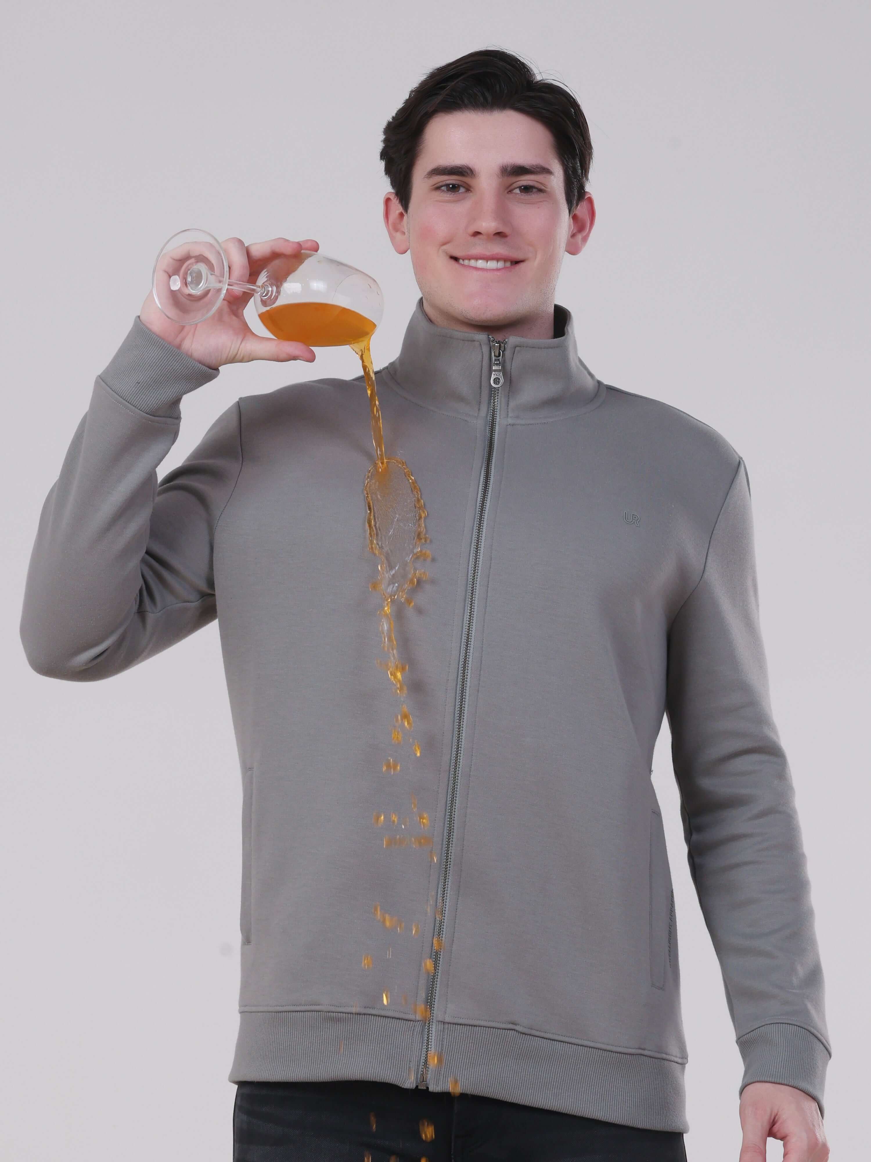Model in a grey zipped jacket demonstrating anti-stain feature while pouring liquid.
