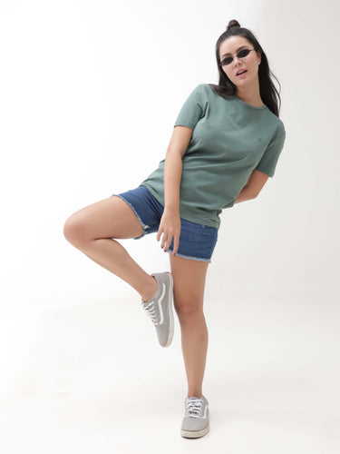 Woman in green Simple Green round-neck t-shirt and denim shorts posing stylishly against a white background