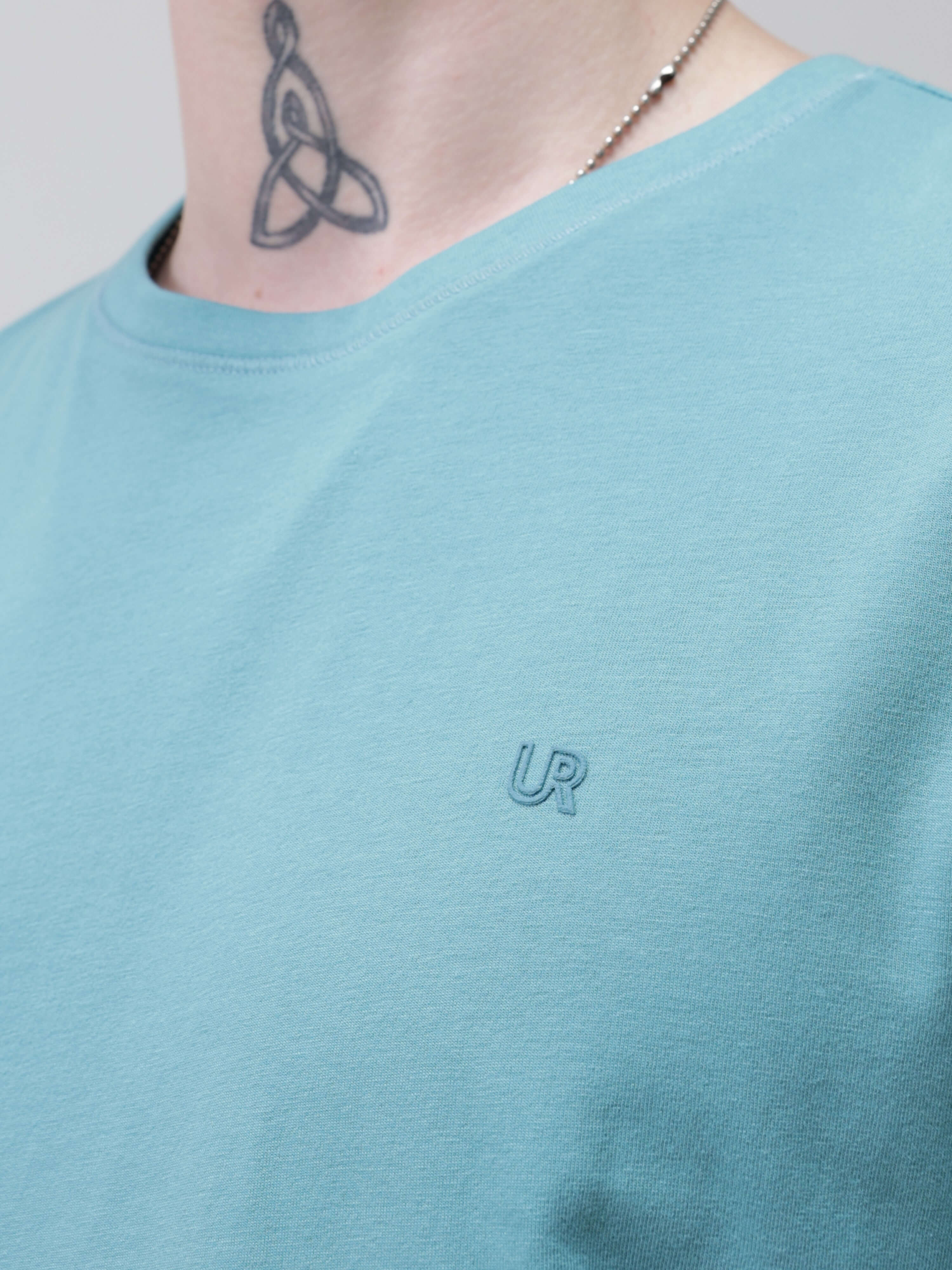 Close-up of a man wearing a light blue round-neck Turms T-shirt, showcasing the brand's logo embroidered on the chest.