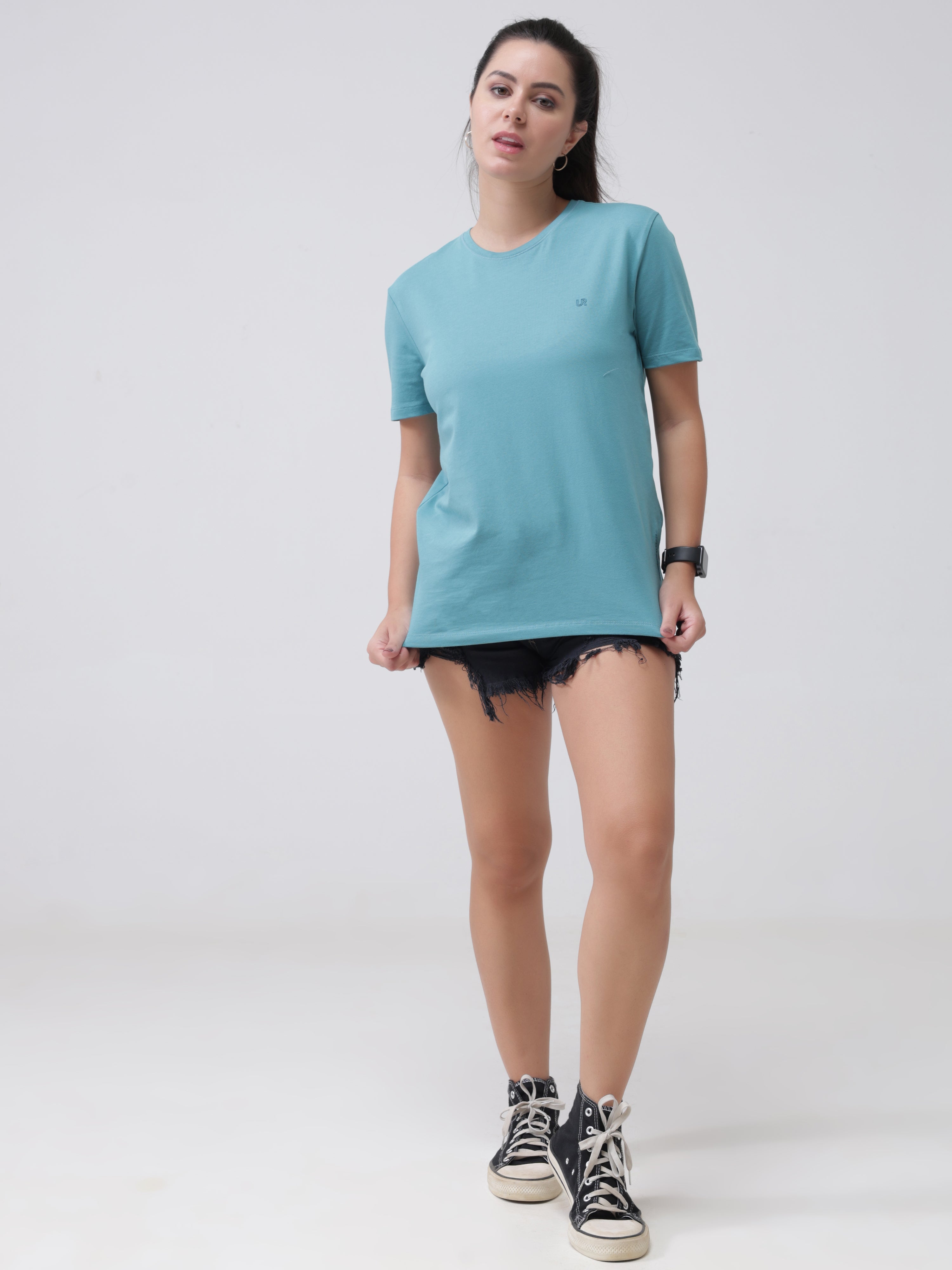 Woman wearing a round-neck turquoise Turms T-shirt with black denim shorts and sneakers, showcasing stylish and stain-proof casual apparel.