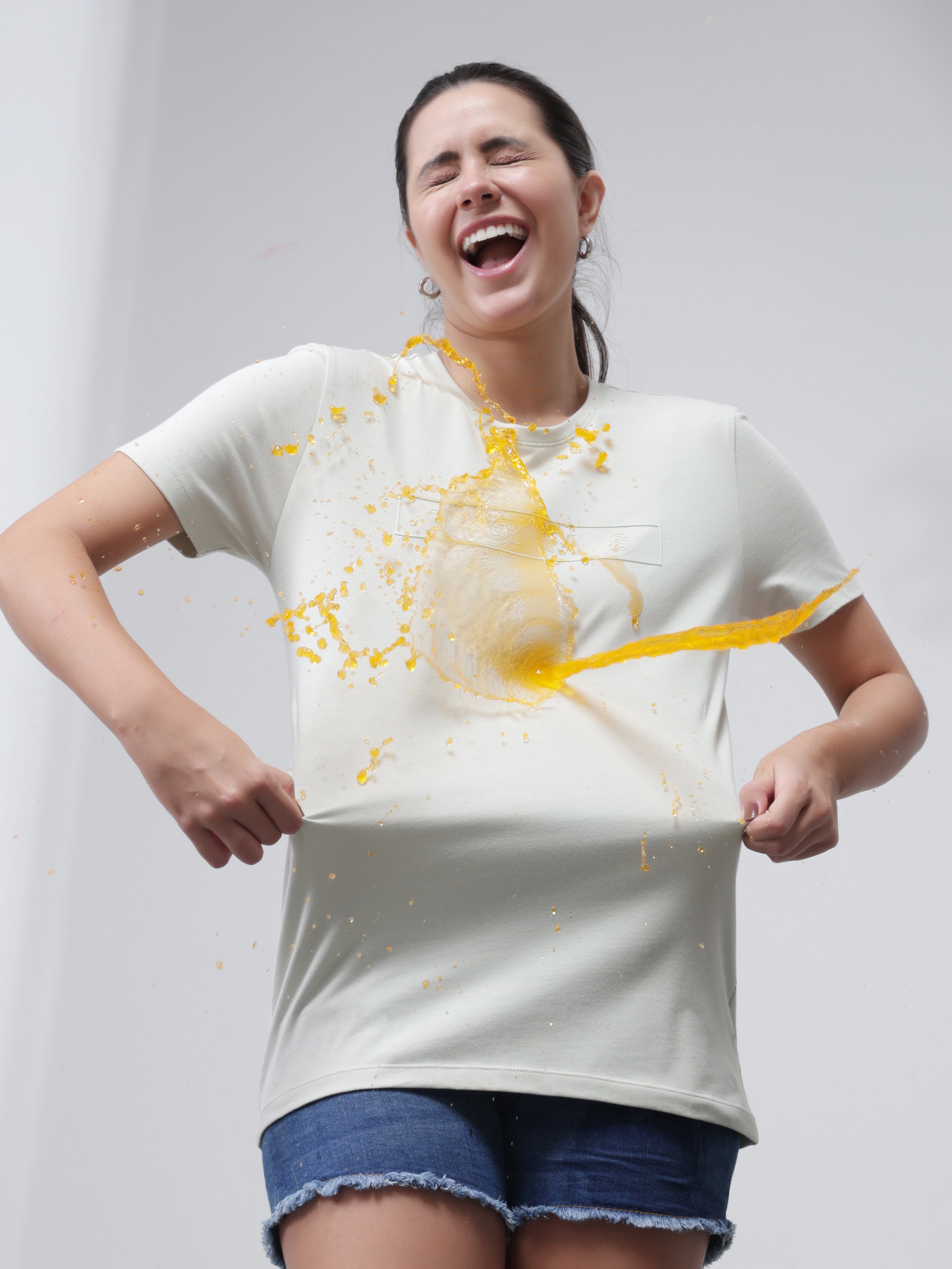 Woman demonstrating stain-repellent Lime Enigma T-shirt by Turms with yellow liquid spill, showcasing anti-stain and anti-odor features.