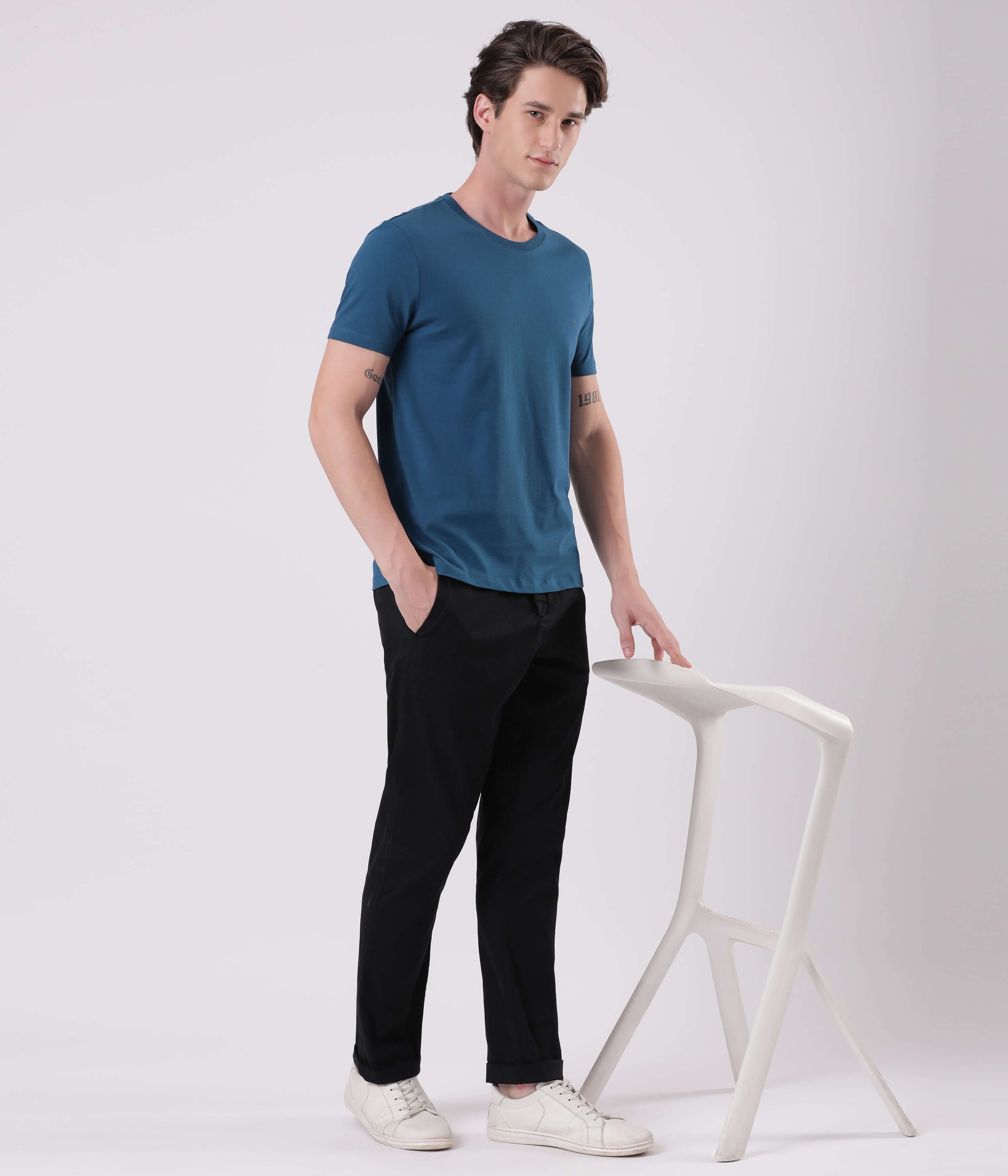 Model wearing Turms Black Sea Chinos in black paired with a teal t-shirt and white sneakers, standing by a minimalist stool.