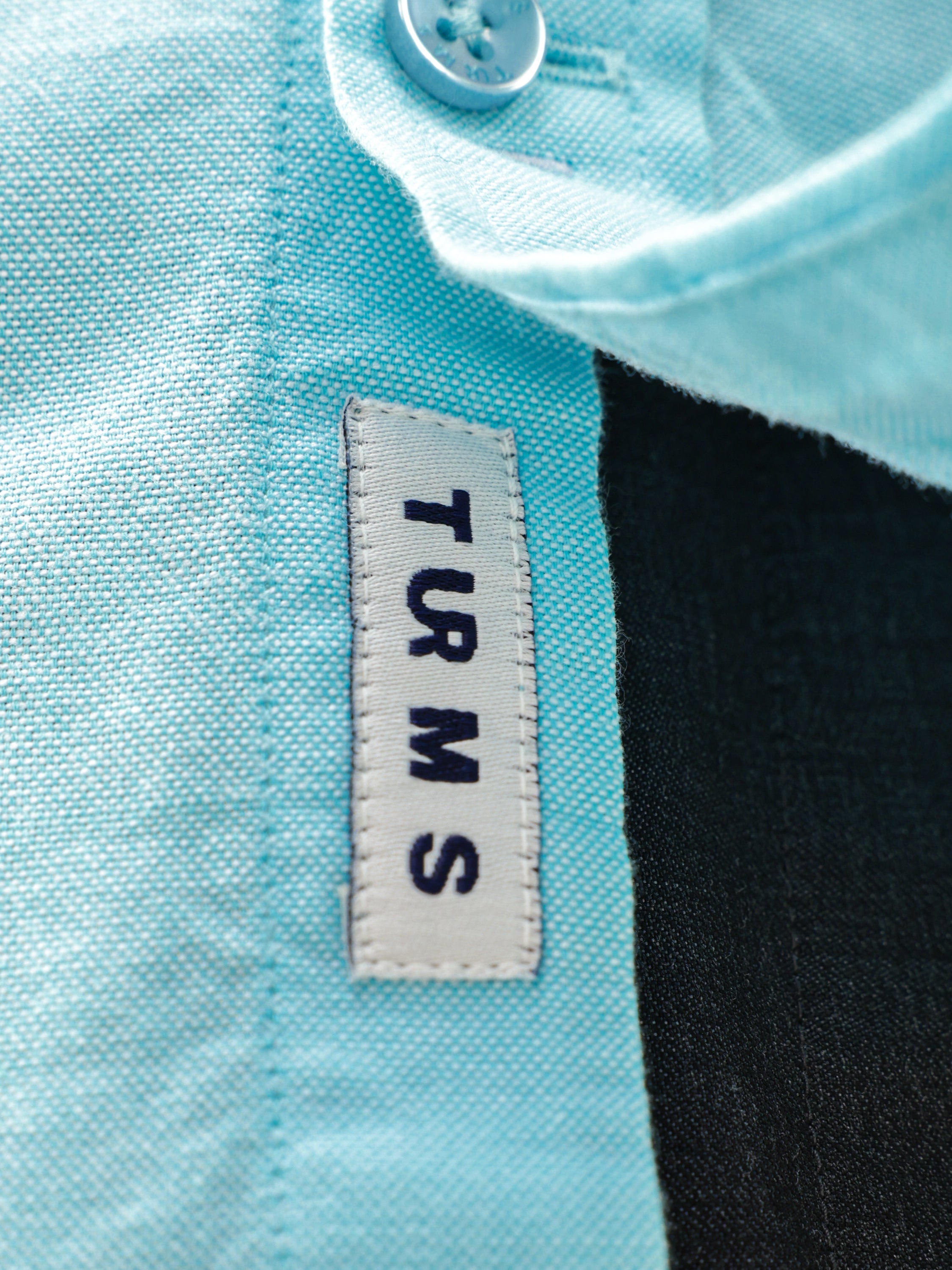 Close-up of Turms tag on a turquoise Oxford shirt, showcasing premium stitching and button detail for menswear.